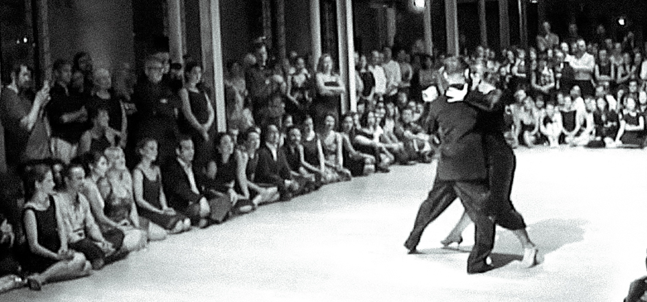 A pair of tango dancer are showing in a milonga hall surrounded by hundred of spectators.