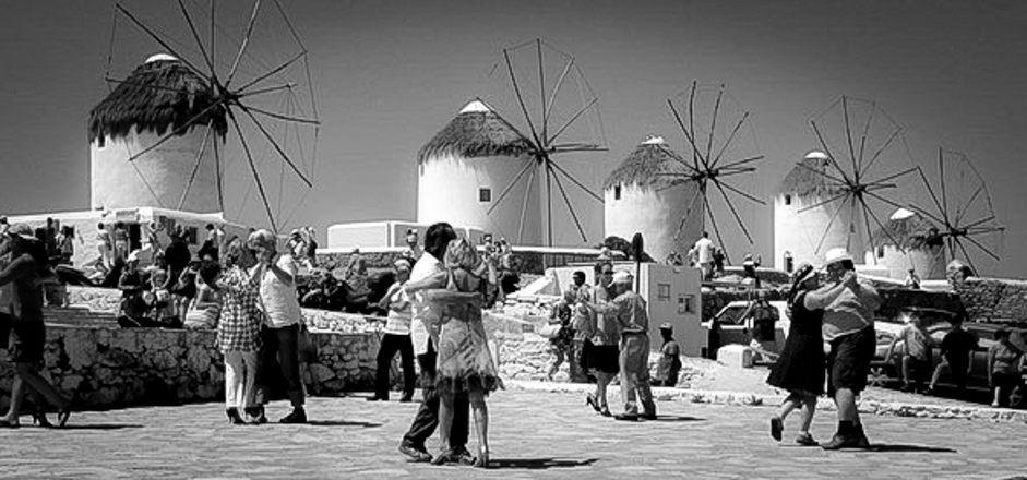 Several tango dancers are dancing outdoor Mykonos with Mykonos Windmills in the background.